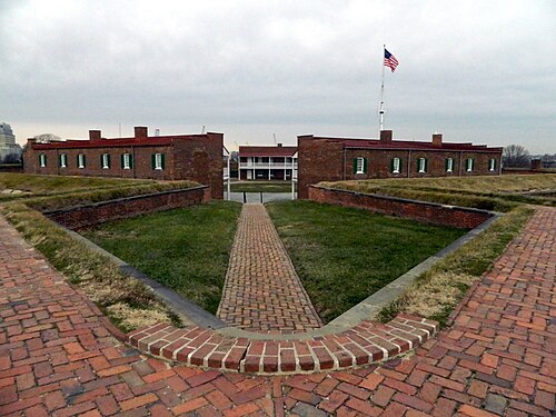 A symmetrical brick path leading between grass covered earthworks to Fort McHenry's brick barracks buildings and a flagpole.