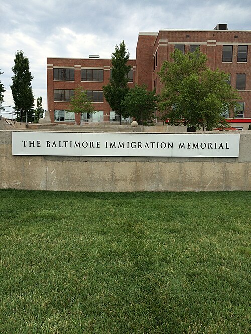 A low granite wall lettered THE BALTIMORE IMMIGRATION MEMORIAL, backed by a lawn, trees and a three storey brick building.