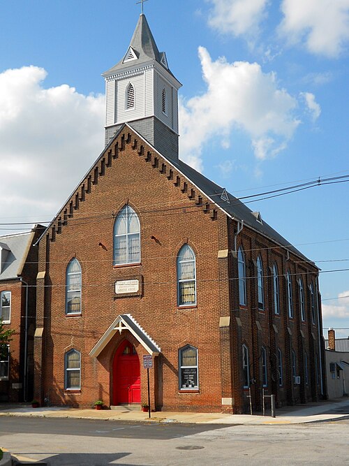 A red brick church with a white wooden steeple and arched window frames, a red door under a small gabled porch, on a Locust Point corner.