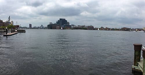 A wide view across the Patapsco River from Locust Point toward the downtown Baltimore skyline, a sailboat and a crane visible on the water.