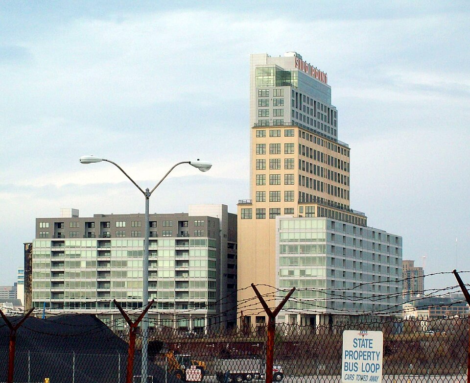 A tan and grey tower with setback upper floors and a rooftop sign reading SILO POINT, rising behind lower glass walled wings, seen from across a fenced rail yard.