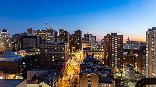 A dusk view south along Cathedral Street from a Mount Vernon rooftop, city rooftops and lit towers toward downtown Baltimore.