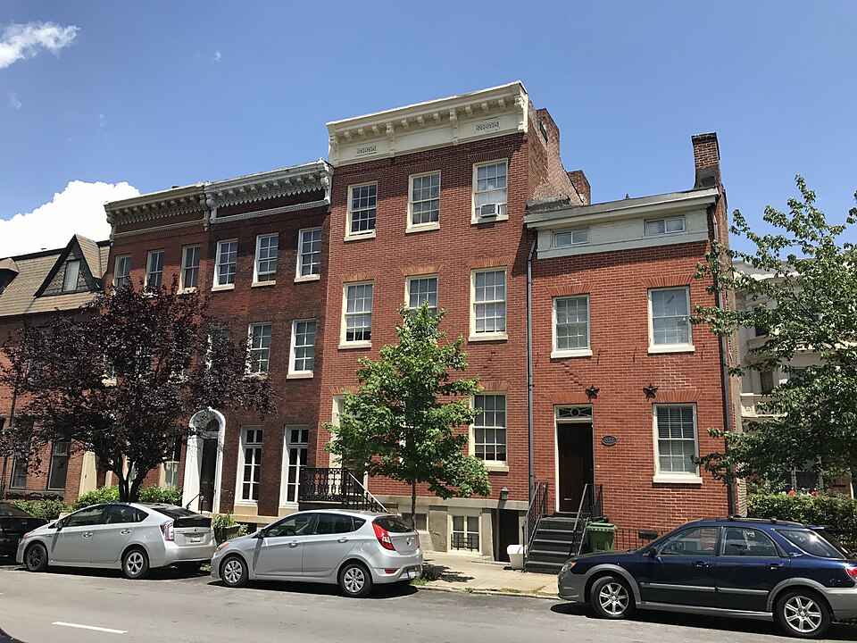 A row of red brick rowhouses on Park Avenue, stone steps and cornices repeating down the block, cars parked along the kerb.