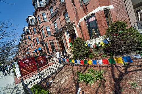 A brownstone row with a shopfront at street level, prayer flags strung along the front railing and pedestrians on the sidewalk.