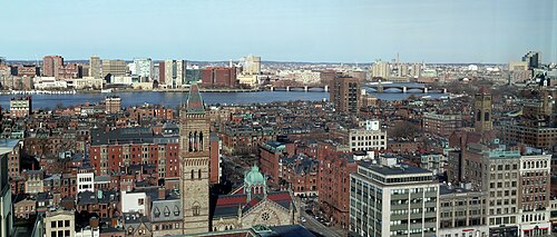 A wide aerial view over Back Bay rooftops and a church tower with a copper roof, the Charles River and the Cambridge skyline beyond.