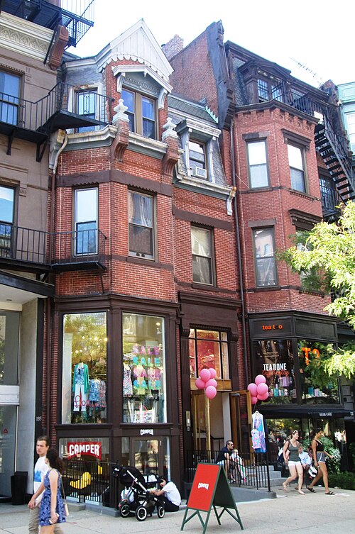 A red brick bowfront between Clarendon and Dartmouth Streets, glass shopfront windows and sale signs at street level under a fire escape and a bay window above.