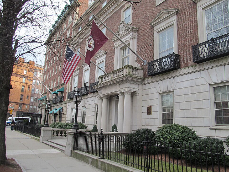 The Harvard Club of Boston's dark red brick clubhouse on Commonwealth Avenue, a columned entrance with iron balconies and a crimson flag above the door.