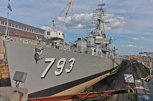 The grey destroyer USS Cassin Young resting in a granite dry dock, its hull number 793 painted large on the bow, a dockside crane and a brick shipyard building behind it.
