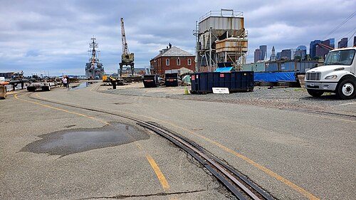 Embedded rail track curving across the paved Navy Yard, a rusted industrial hopper and the Shipyard Galley building beside it, the destroyer moored at the pier on the left and the downtown Boston skyline behind.