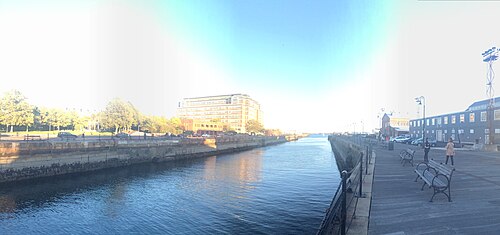 A wide waterfront promenade along a narrow stone lined channel, a large brick building at the far end, benches along the boardwalk and a smaller shingled building with a lighting rig on the opposite bank.