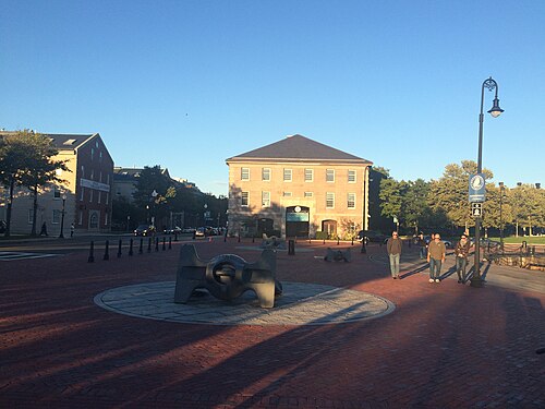 A brick paved plaza with two large bronze ship's anchors mounted as sculpture, a granite lodge building behind, and a street lamp with a Charlestown neighborhood marker.