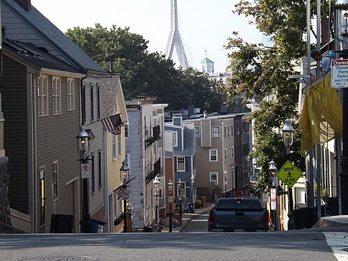 A steep residential street of painted wood houses running downhill, string lights and street lamps along the sidewalk, the cables and towers of the Zakim Bridge rising above the rooftops at the bottom.