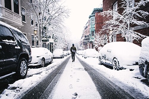 A snow covered residential street between red brick and painted wood rowhouses, a bowfront iron balcony on the right, a person walking up the unplowed middle of the road past snow covered parked cars.