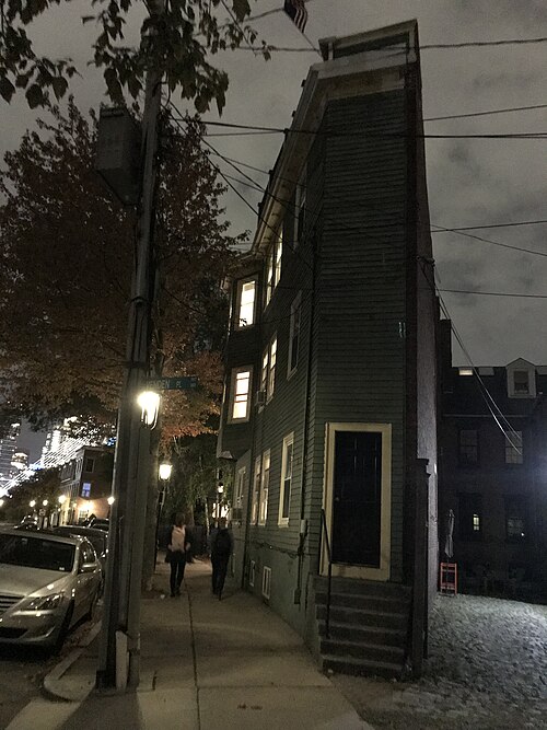 A narrow three storey green clapboard house wedged beside a taller brick building at a street corner, photographed after dark with its windows lit and two people walking past on the sidewalk.