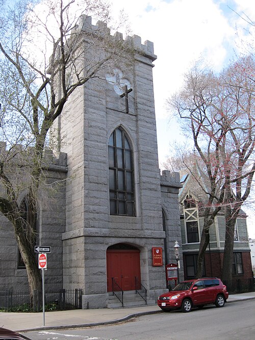A grey stone church tower with battlement parapets and pointed Gothic windows, a wood frame Victorian house and parked cars along the sidewalk beside it.