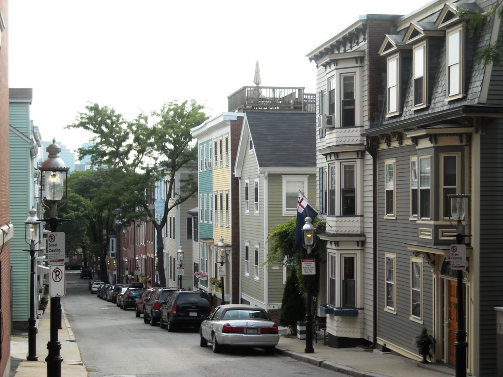 A steep asphalt street lined with painted wood clapboard and brick rowhouses, bay windows and a rooftop deck visible, cars parked nose to tail down the hill and the downtown skyline showing through the trees at the bottom.