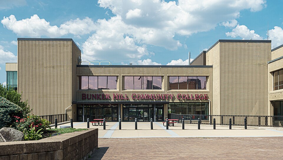 The main entrance plaza of Bunker Hill Community College, a low tan brick building with fluted end towers and the college name in large letters over the glass doors, bollards and picnic tables on the brick forecourt.