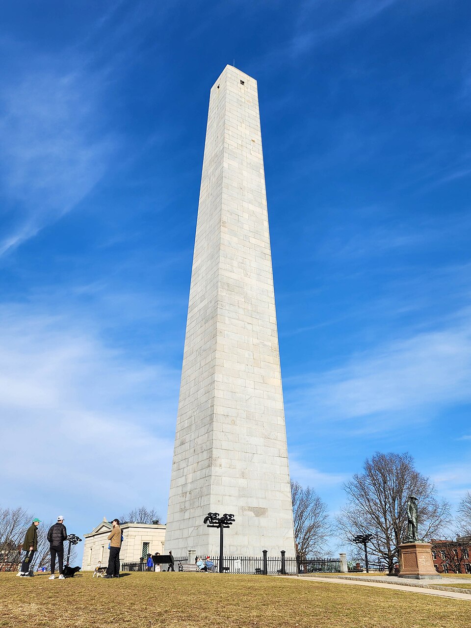 The granite obelisk of the Bunker Hill Monument seen from the base of the lawn, an iron fence around it, a stone lodge building to one side and a bronze statue on a pedestal to the other, a few visitors and a dog on the grass.