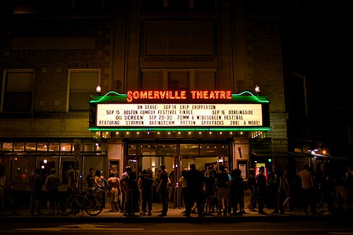 The Somerville Theatre marquee lit in red neon at night, a crowd on the sidewalk under the glass doored entrance.