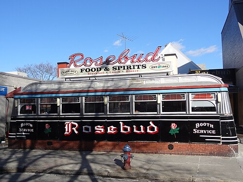 The Rosebud, a chrome streamliner diner car with a red neon sign and black lower panels, on a brick sidewalk with a fire hydrant out front.