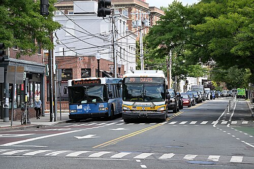 Two MBTA buses stopped nose to tail on College Avenue, brick storefronts and overhead wires along the tree lined street.
