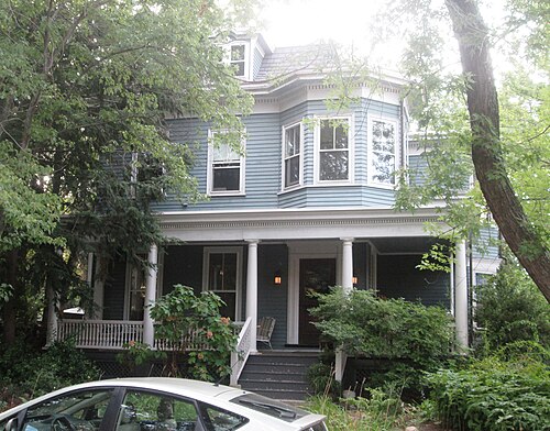 A blue two and a half storey wood frame house with a wraparound porch, white columns and a bay window, set behind trees.