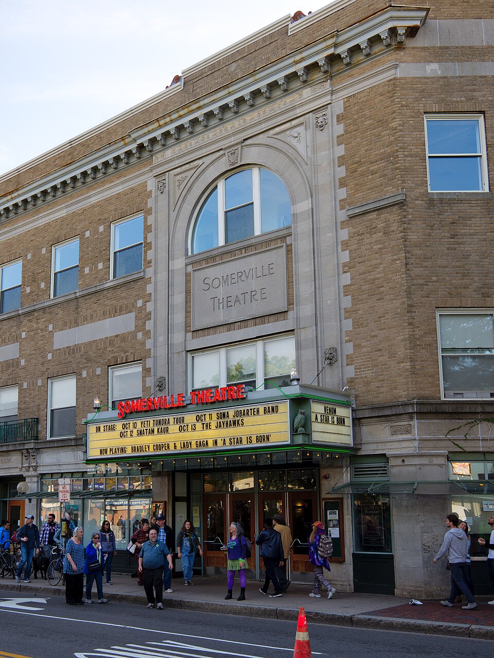 The Somerville Theatre entrance at Davis Square, an arched stone window over the marquee, glass doors under a green awning, and pedestrians on the sidewalk.