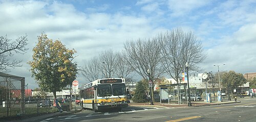 A yellow and white MBTA bus signed 18 Ashmont waits at a paved busway beside bare trees, storefronts and a round public clock behind it.