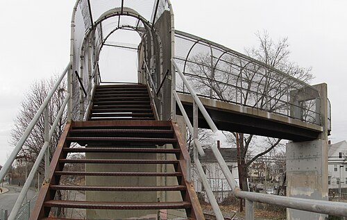 A rusted steel footbridge with a mesh cage over the Fairmount Line, houses visible through the bare trees beyond.