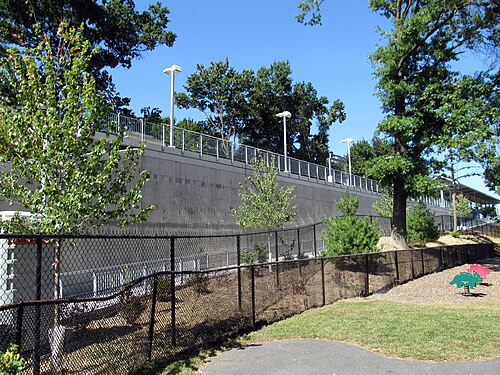 A concrete retaining wall and fenced ramp leading up to the inbound platform at Four Corners and Geneva station, a small park and playground equipment below.