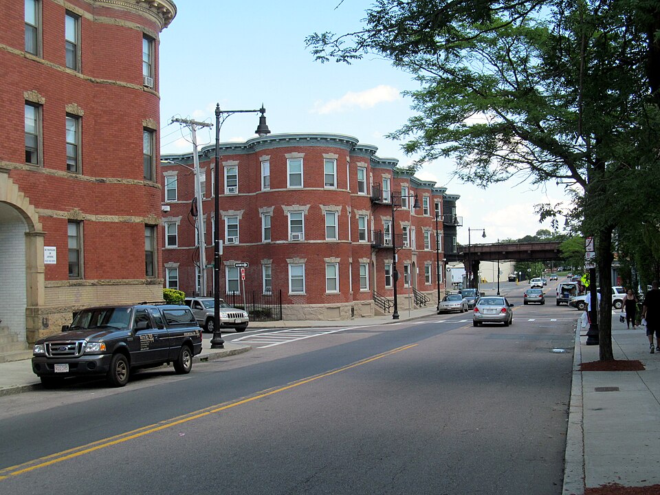 Brick three storey apartment buildings on Geneva Avenue near Fields Corner, bow fronted with white window trim, parked cars along the curb and a street lamp in front.