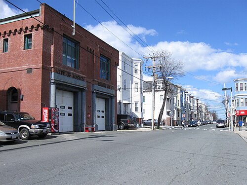 A red brick fire station with two arched engine bay doors labeled Engine Co. 5, wood frame triple deckers down the block behind it.