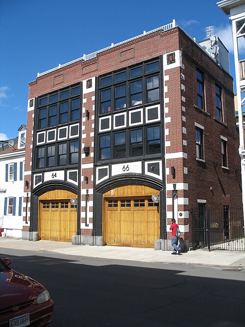 A converted red brick firehouse on Marion Street, two arched wooden garage doors and white quoin trim, a wood sided rowhouse next door.