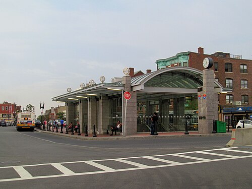 The MBTA Blue Line's Maverick Station, a curved glass and granite headhouse beside an older brick commercial building with a corner clock.