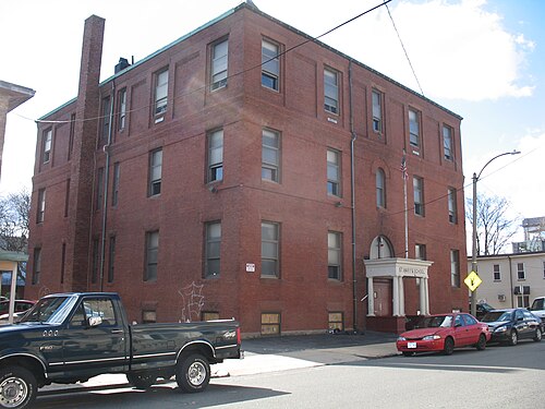 A red brick school building on a raised basement, three rows of tall windows and a white columned entrance portico reading St. Mary's School, cars parked along the curb.
