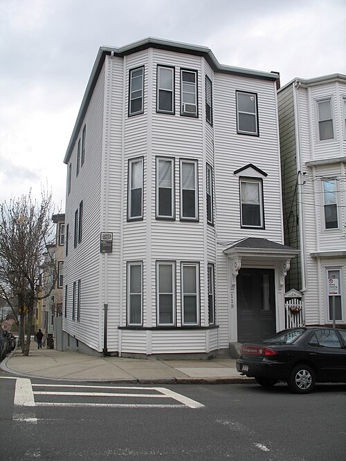 A three storey white sided corner house, bay windows stacked up its street facing corner, a black door under a gabled hood, a parked car at the curb.