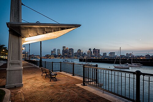 A lit park pavilion and railed promenade at Piers Park, sailboats moored on the harbor and downtown Boston's towers across the water at dusk.