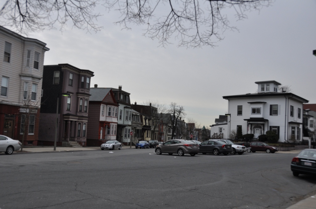 A wood frame triple decker street meeting at a junction on Eagle Hill, bay windows and a mansard tower on the corner house, cars parked along the curb. The kind of Eagle Hill street the Equalizer used a house on, not Falcon Street itself.