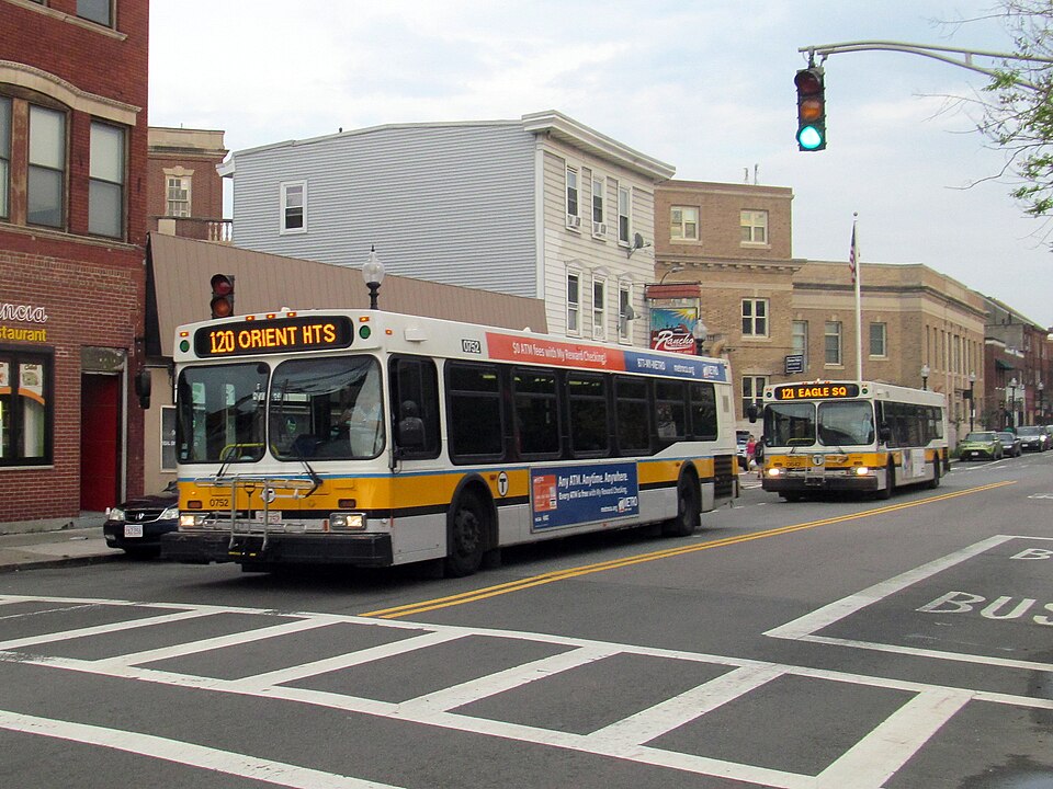 Two MBTA buses stopped on Meridian Street near a corner restaurant sign, brick and wood sided buildings behind under a traffic signal. The kind of East Boston street near Maverick Square that Equalizer 2 closed for its car chase, not Orleans Street itself.