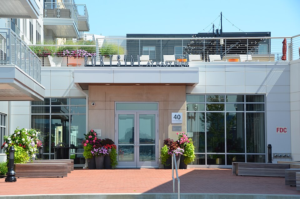 The Portside at East Pier building's own entrance, a glass door under the development's sign, potted flowers and a wood bench on the brick plaza in front, the numbered plaque reading 40 East Pier Drive beside the door.
