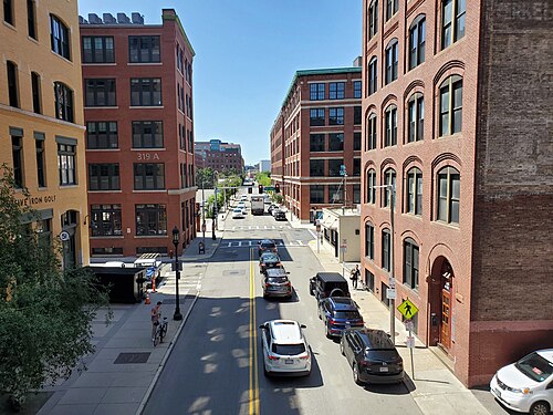An elevated view down A Street between red and yellow brick warehouse buildings, a 319 A address number, parked cars and a cyclist on the roadway.