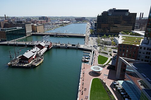 An aerial view of Fort Point Channel, its bridges, the Boston Tea Party Ships and Museum on the water, and brick warehouses along both banks.