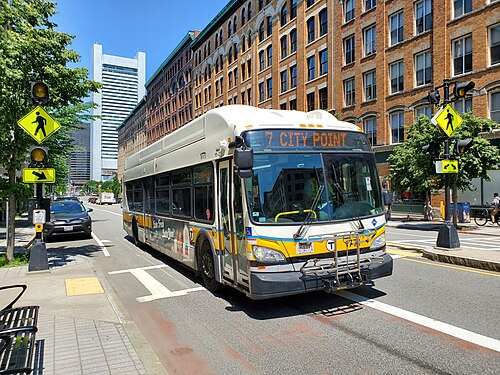 An MBTA route 7 bus passing a brick warehouse with arched window openings running the length of the block.