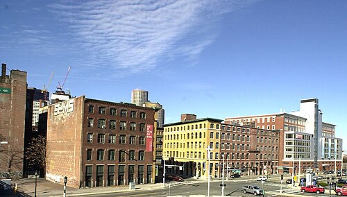 An elevated view of a brick warehouse corner with a faded painted sign on its blank wall, cranes and downtown towers in the distance.