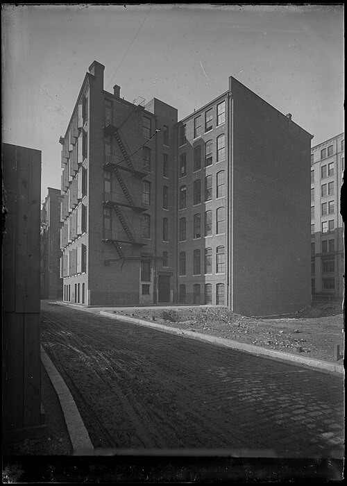 The rear of a six storey brick warehouse on Melcher Street, cast iron fire escapes climbing the wall above an unpaved lot, photographed for the Boston Wharf Company.