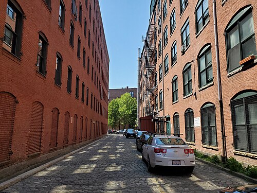 A narrow cobblestone alley between two red brick warehouses, cast iron fire escapes and arched window openings on both sides, parked cars along the curb.