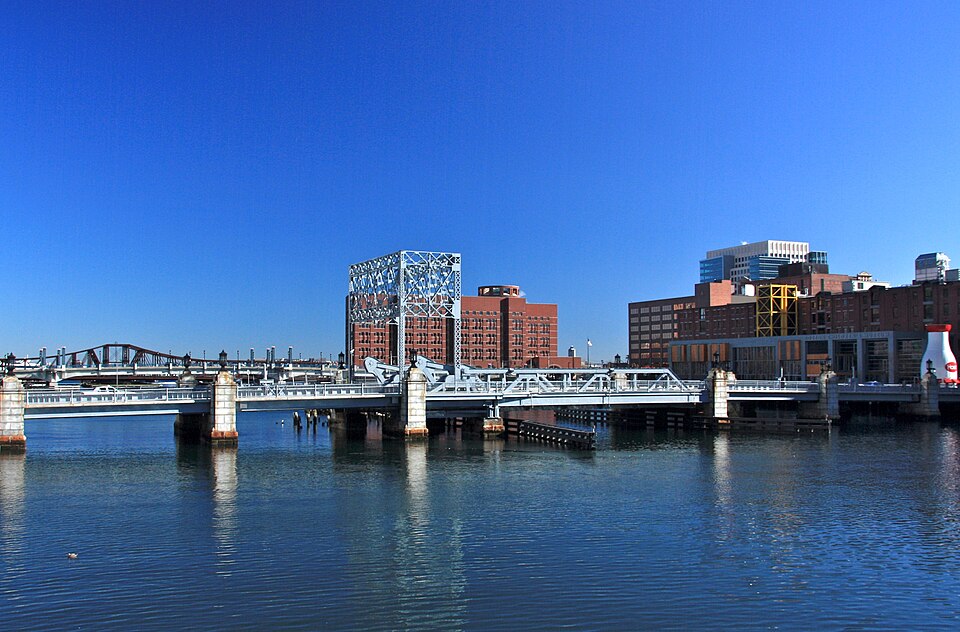 The steel bascule bridge over Fort Point Channel, its lattice tower raised beside the roadway, brick warehouses and the Hood Milk Bottle visible on the far bank.