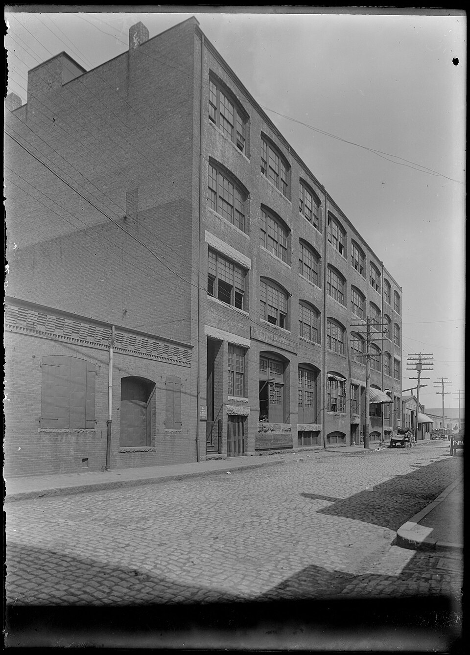 A five storey brick warehouse on a cobblestone stretch of Farnsworth Street, telephone poles and wires strung along the curb, photographed for a Boston Wharf Company real estate survey.