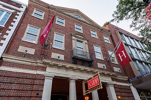 The Harvard Cooperative Society's red brick Georgian facade, a maroon flag and an H banner either side of the pedimented doorway, THE COOP sign over the columned entrance.
