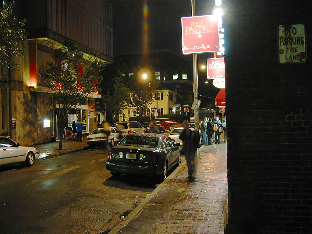 A brick sidewalk on a Harvard Square side street at night, parked cars and a few pedestrians under a red PHO PASTEUR sign and a NO PARKING FIRE LANE notice on the corner.